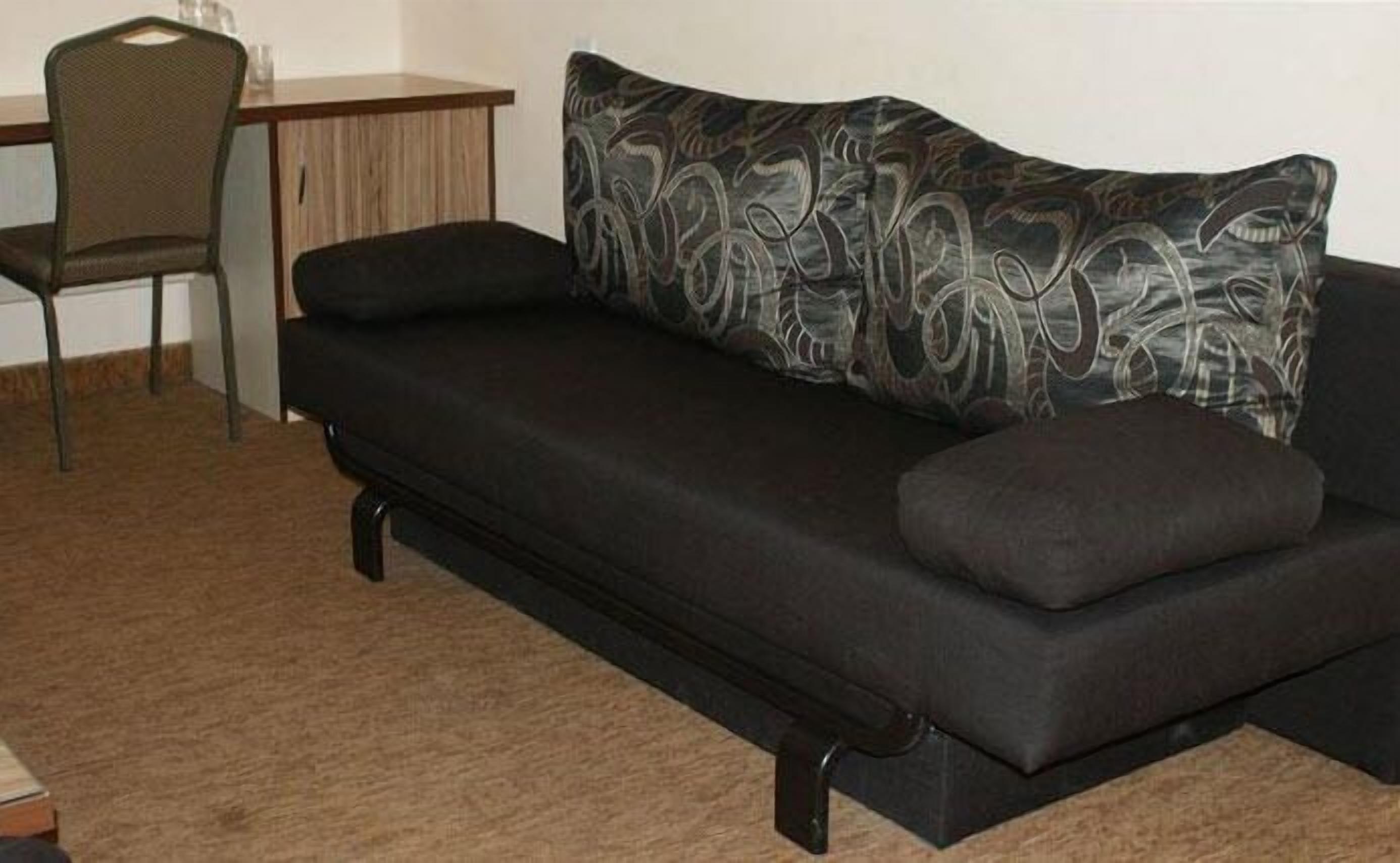 Un sofá negro con cojines estampados en una habitación con un escritorio de madera y una silla.