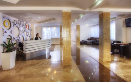 A modern hotel lobby with a reception desk, seating area, and a large TV screen.