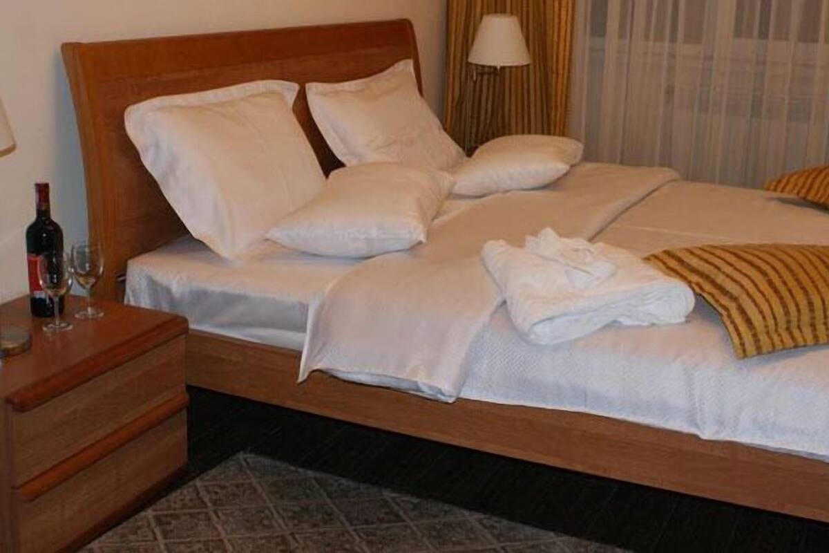 A neatly made bed with white linens and pillows, a wooden headboard, a bedside table with a bottle and glasses, and a patterned rug.