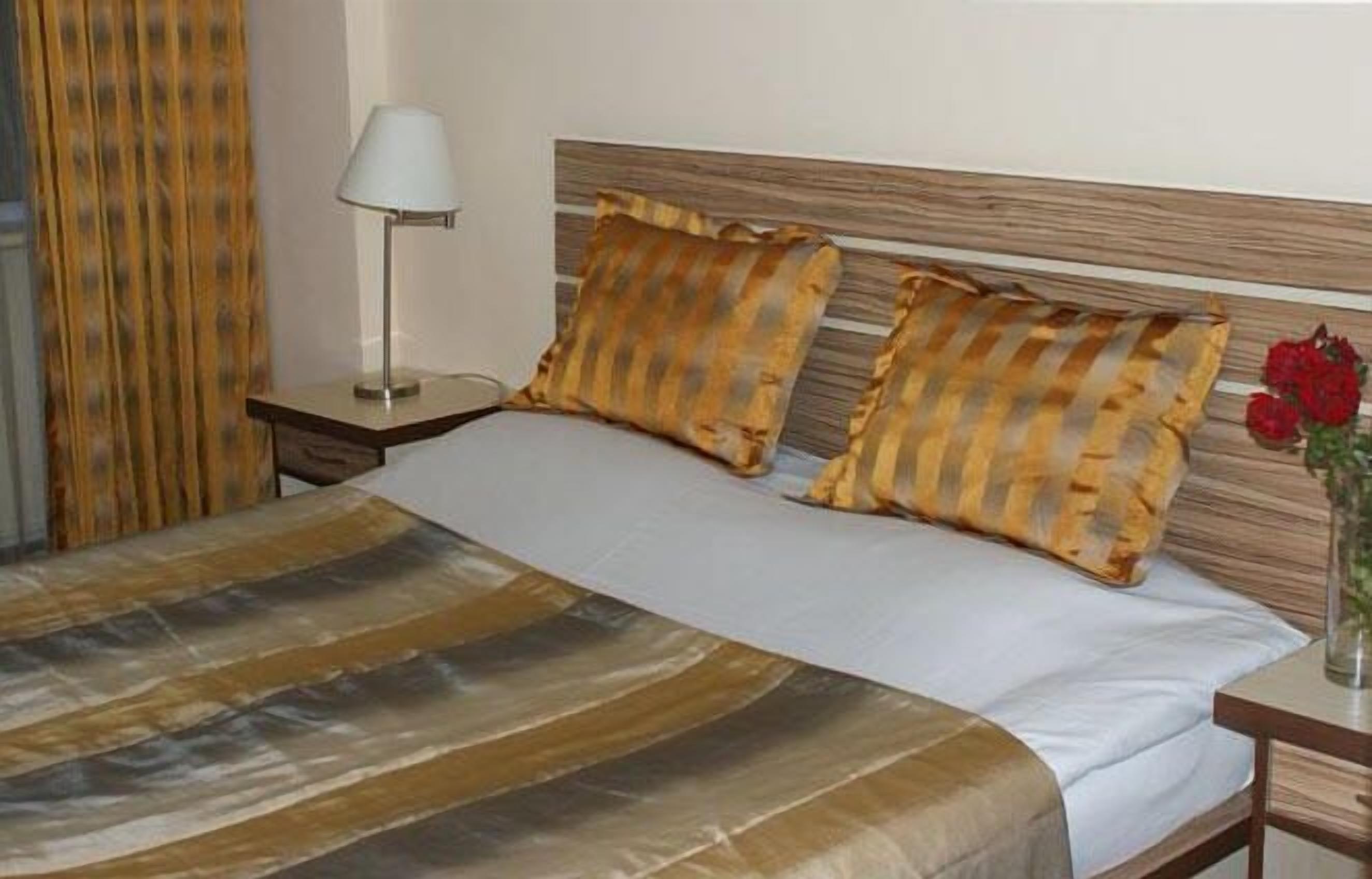 Una cama bien hecha con almohadas a rayas, una mesita de noche con una lámpara y un jarrón con flores.