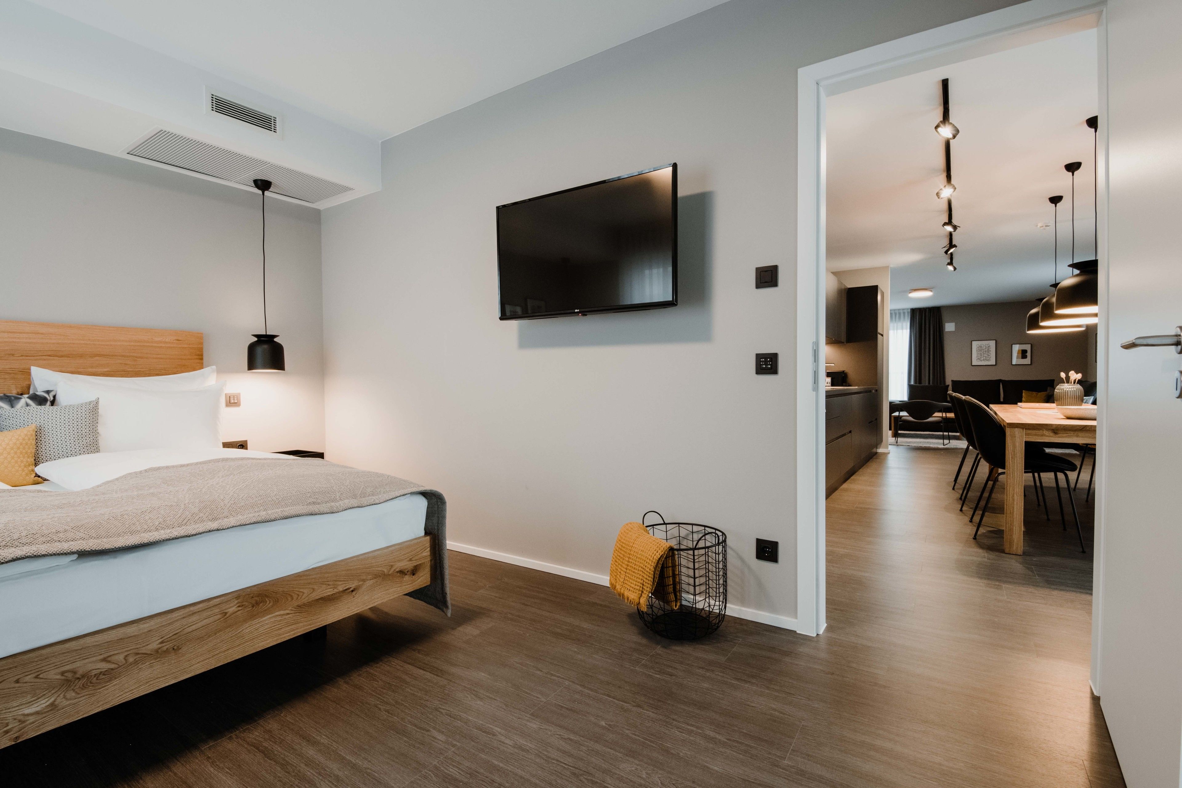 A modern hotel room with a wooden bed, a flat-screen TV mounted on the wall, and a dining area visible through an open door.