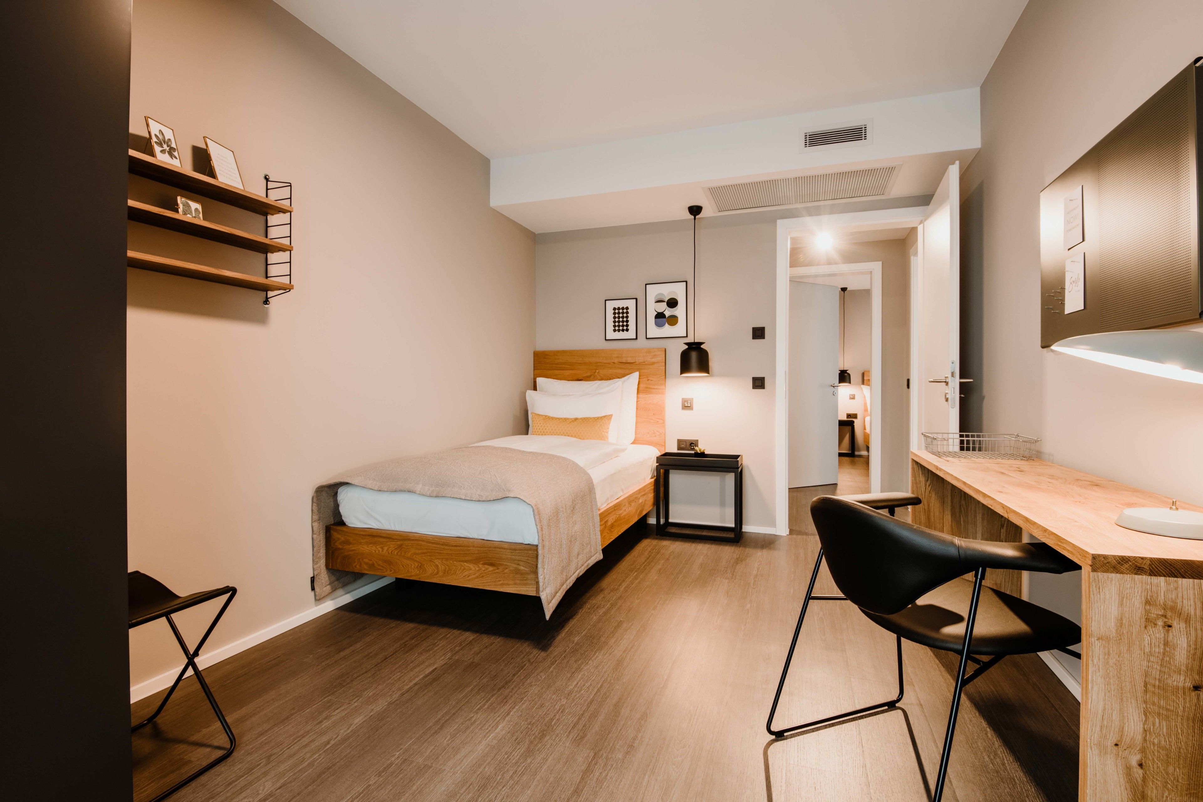 Een moderne slaapkamer met een houten bed, een bureau met stoel, een plank met foto’s en een spiegel aan de muur.