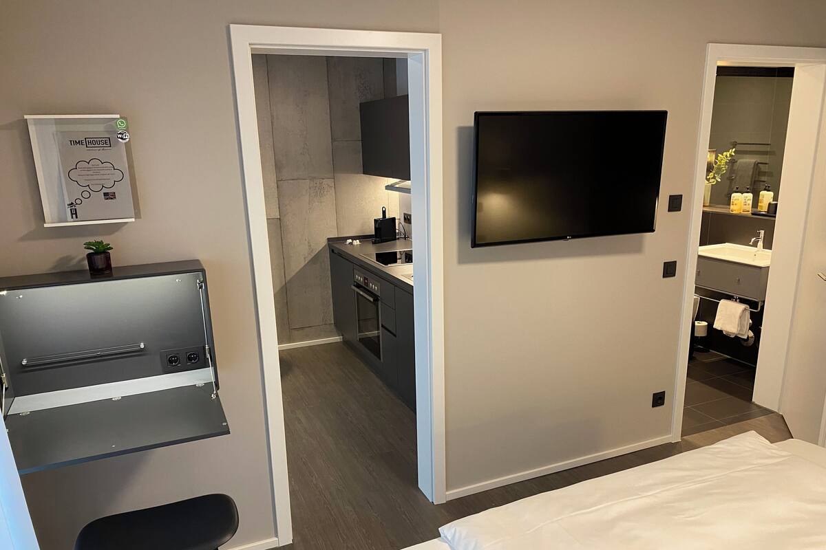 Una habitación de hotel moderna con cama, escritorio con silla, televisor montado en la pared y kitchenette.