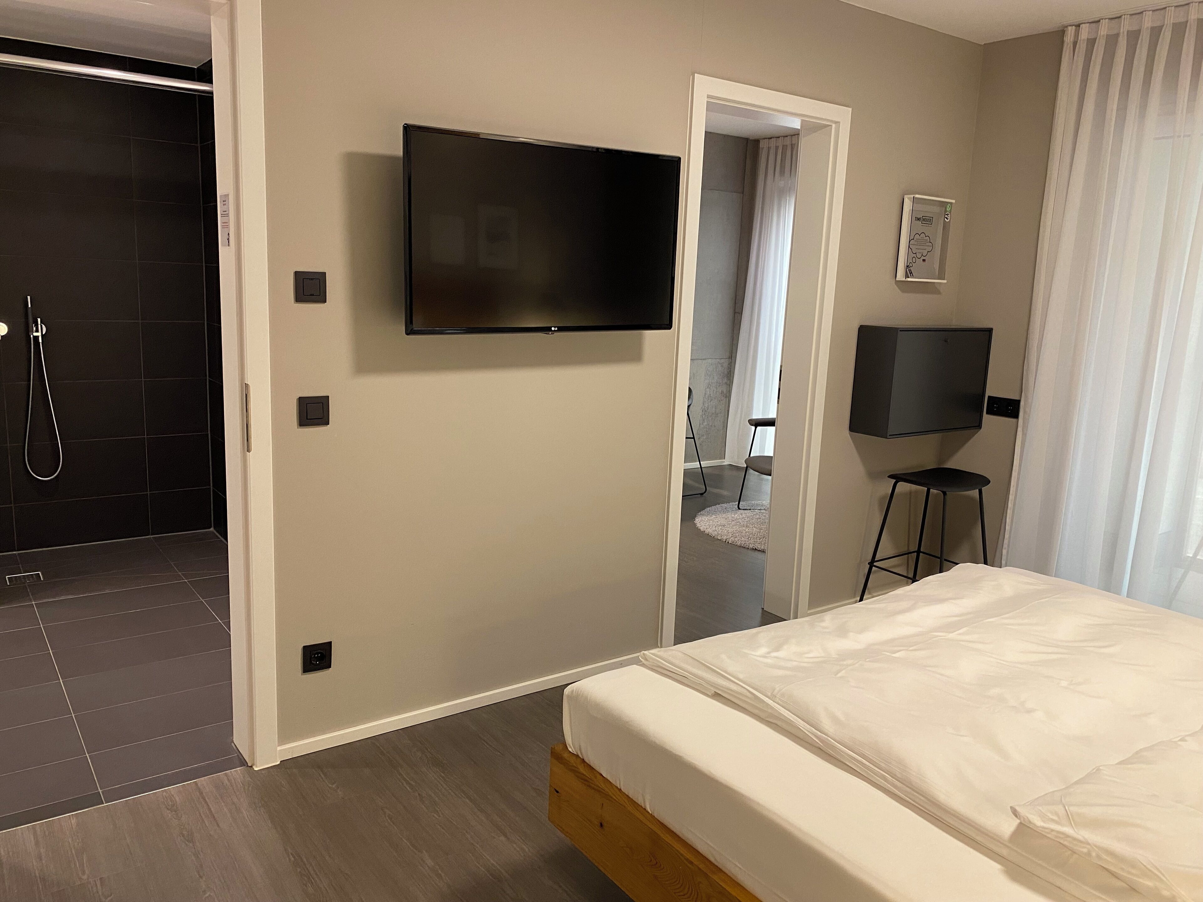 A hotel room with a bed, a TV mounted on the wall, a shower area, and a door leading to a bathroom.