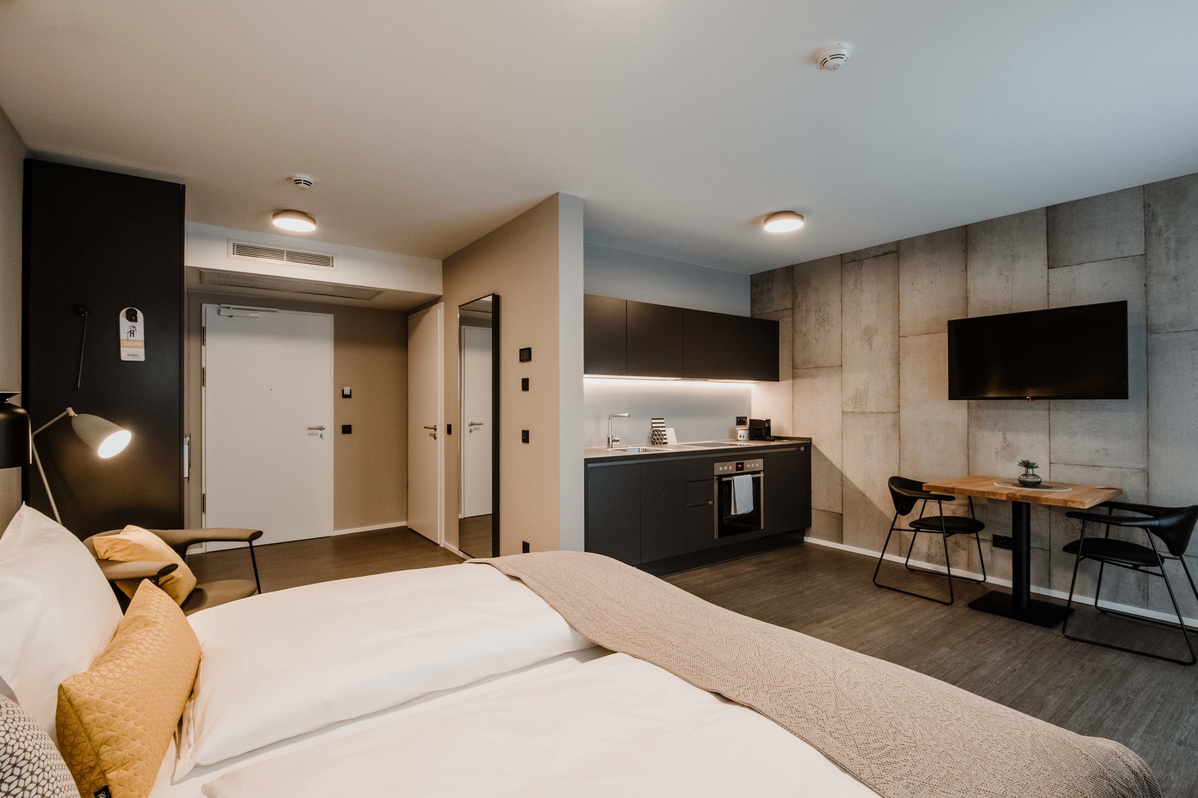 A modern hotel room with a bed, bedside table, lamp, and a kitchenette area with a TV and seating.