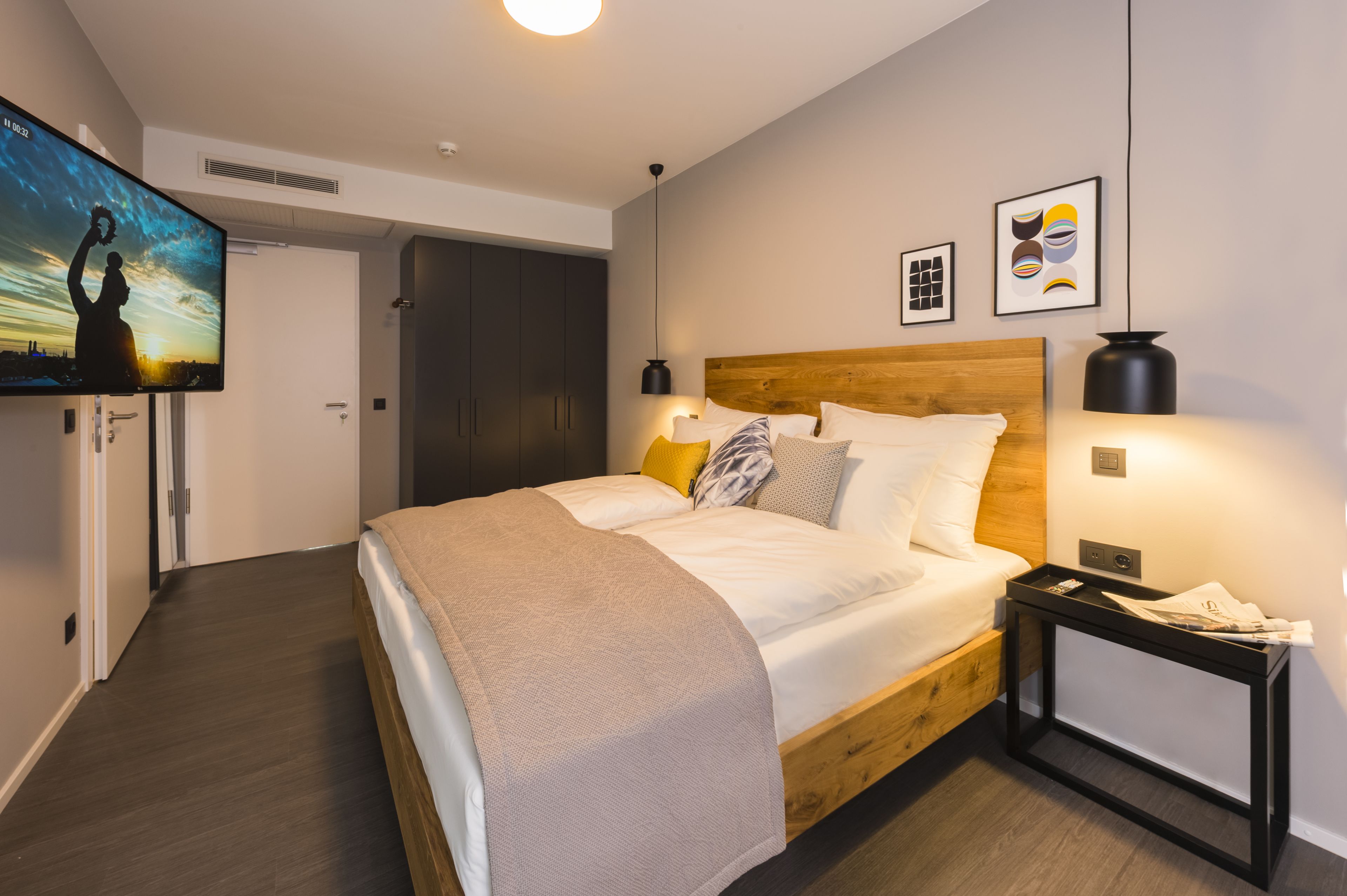 Um quarto moderno com uma cama de madeira, uma mesa de cabeceira, televisão montada na parede e obras de arte nas paredes.