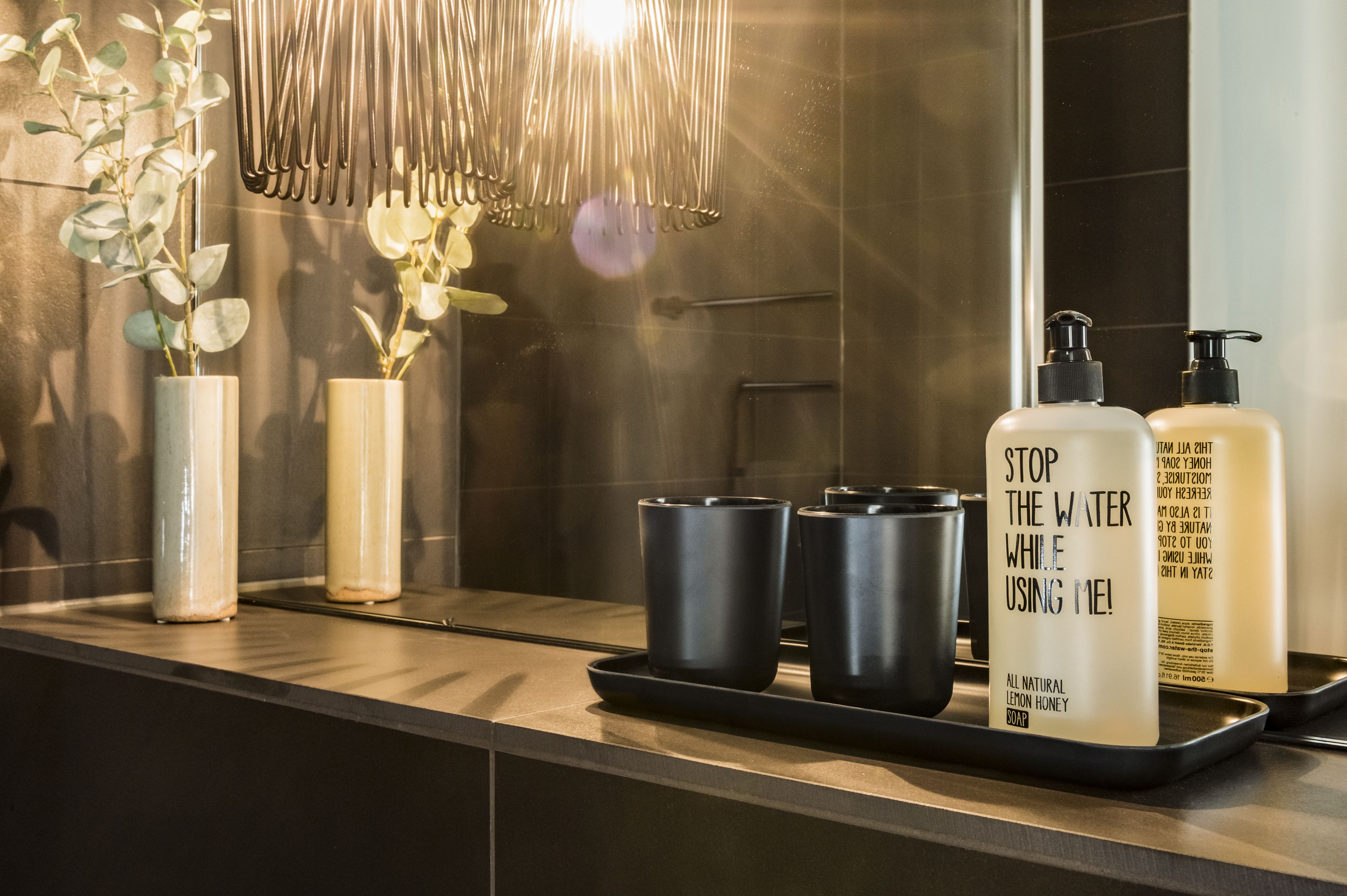A bathroom vanity with a mirror, two vases with plants, a tray with two black cups, and two bottles of soap.