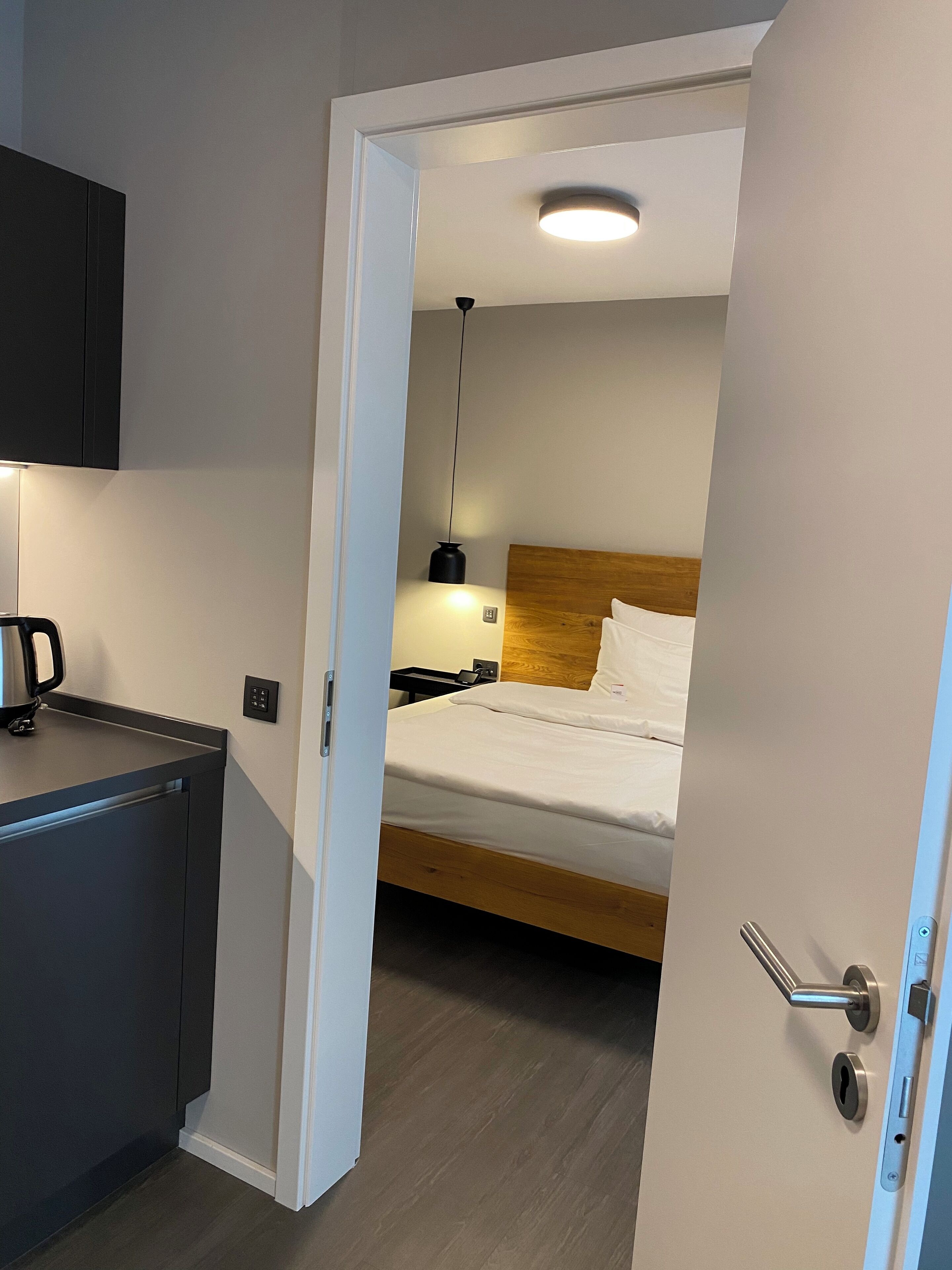 A hotel room with a bed, bedside table, and a small kitchenette area.