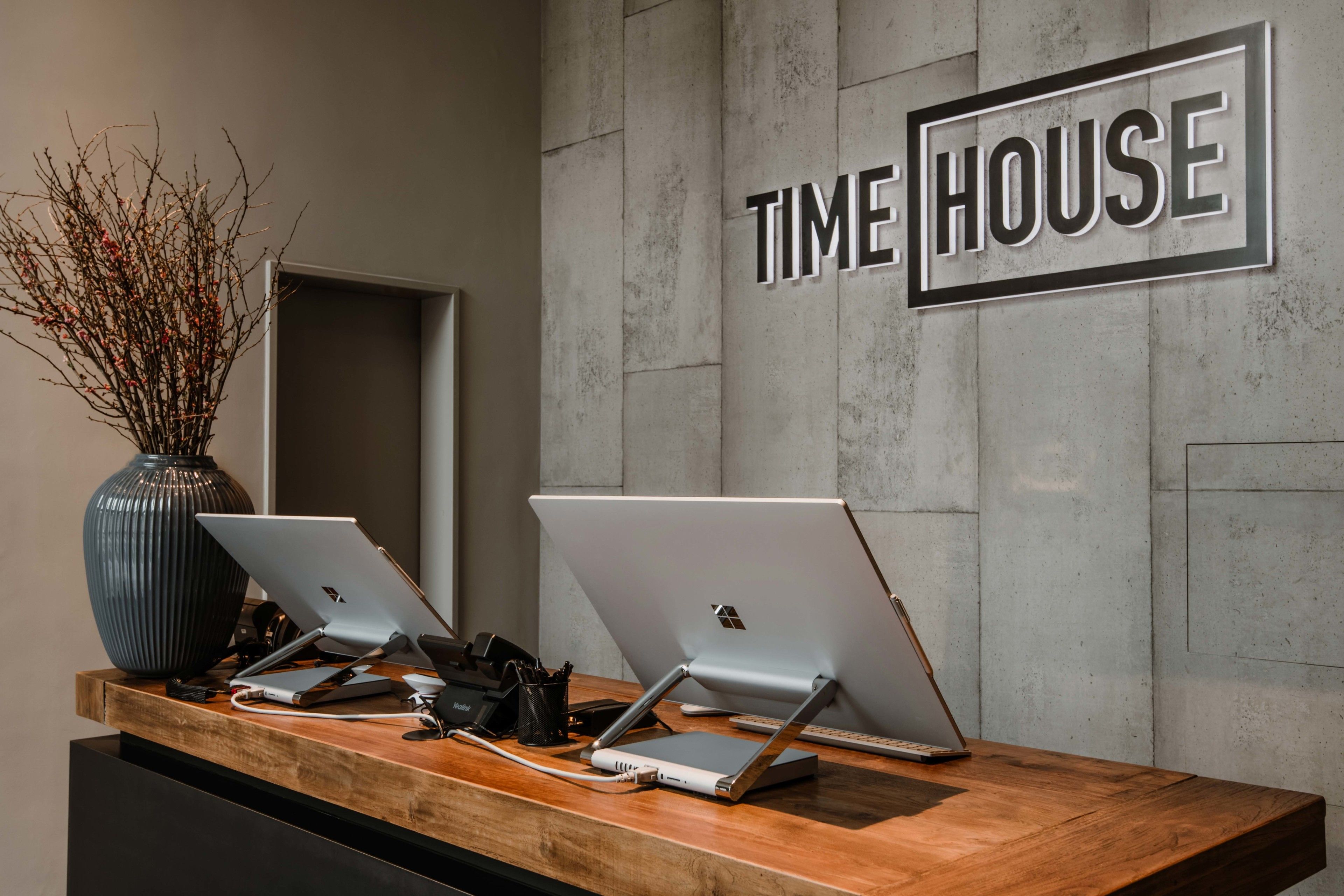 Una recepción con dos computadoras portátiles, un florero con ramas y un letrero en la pared que dice 'TIME HOUSE'.