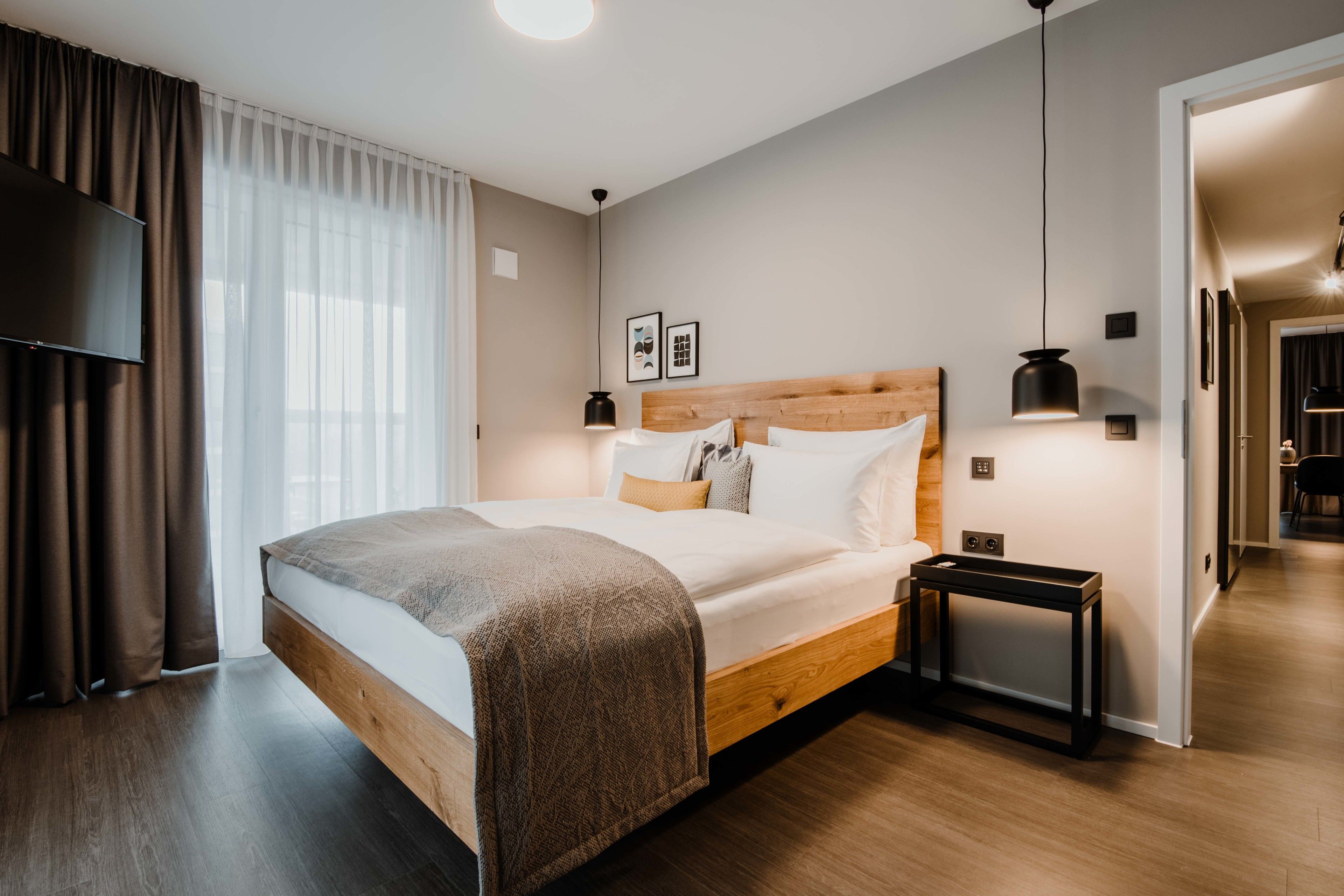 A modern bedroom with a wooden bed, a bedside table, a TV, and a view of a hallway.