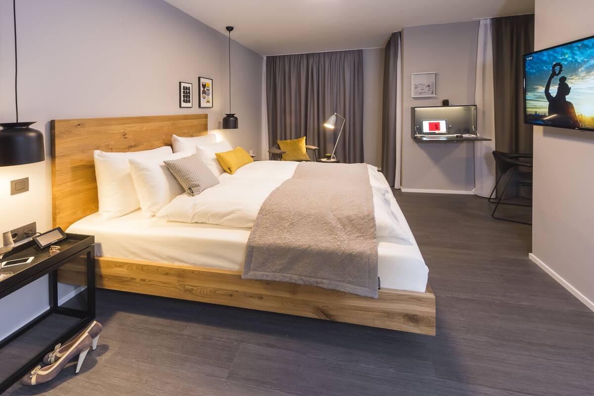 Una habitación de hotel moderna con una cama de madera, un escritorio con una computadora portátil, un televisor y una pequeña zona de estar.