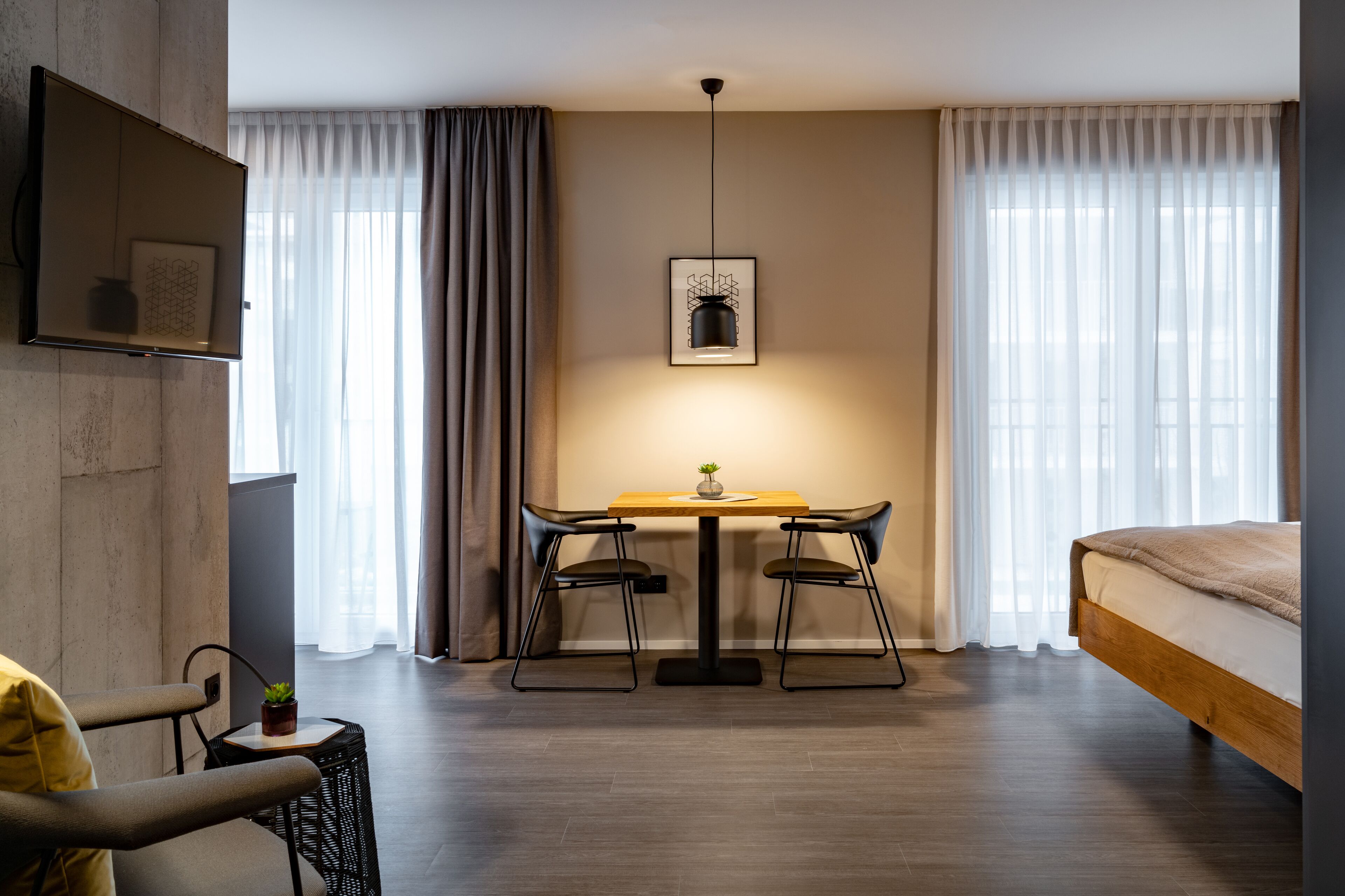 Quarto de hotel moderno com piso de madeira, uma cama, uma mesa de jantar com duas cadeiras, uma mesinha lateral com uma planta e uma TV fixada na parede.