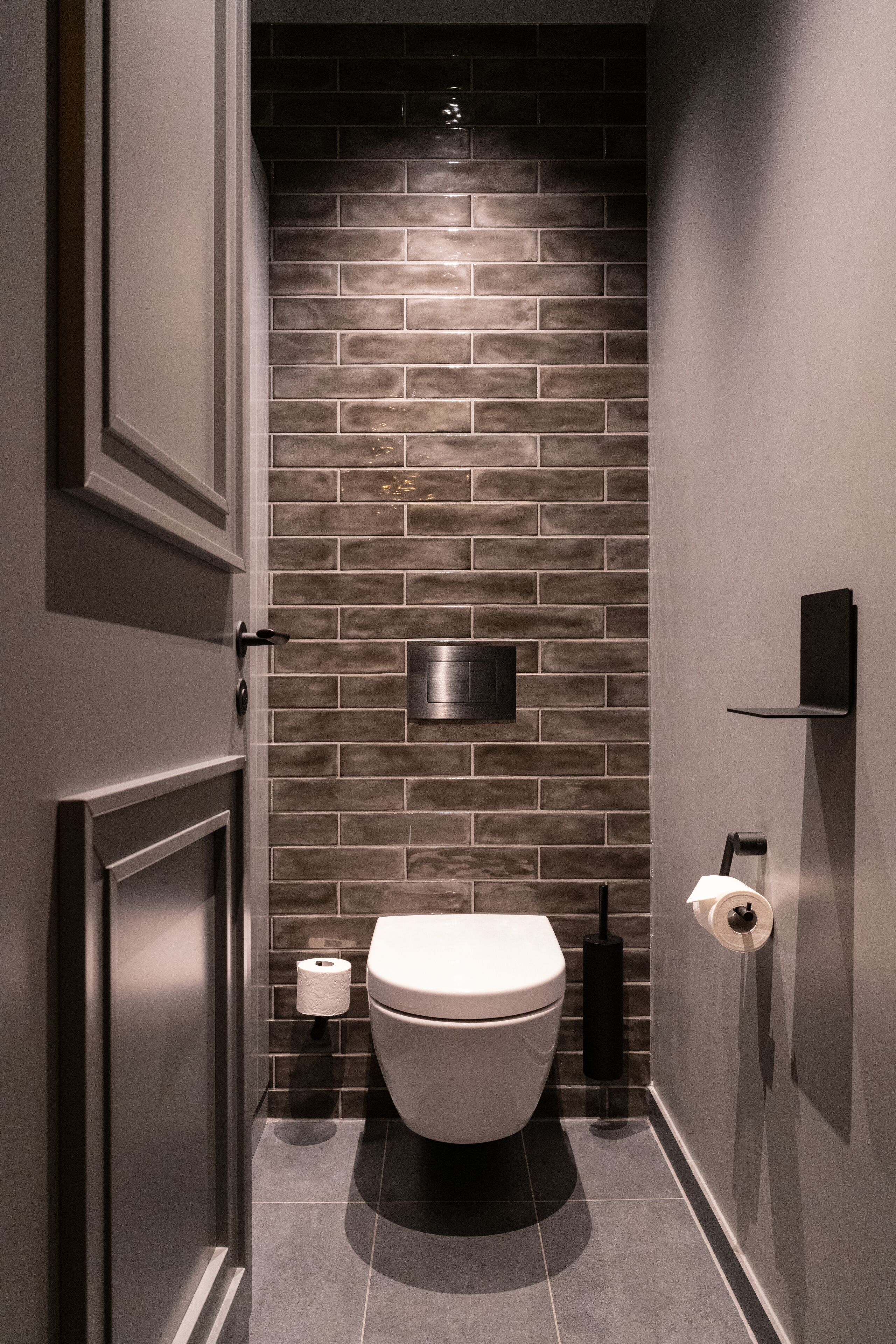 Un baño moderno con inodoro blanco, pared de ladrillo oscuro y un estante pequeño con un objeto negro.