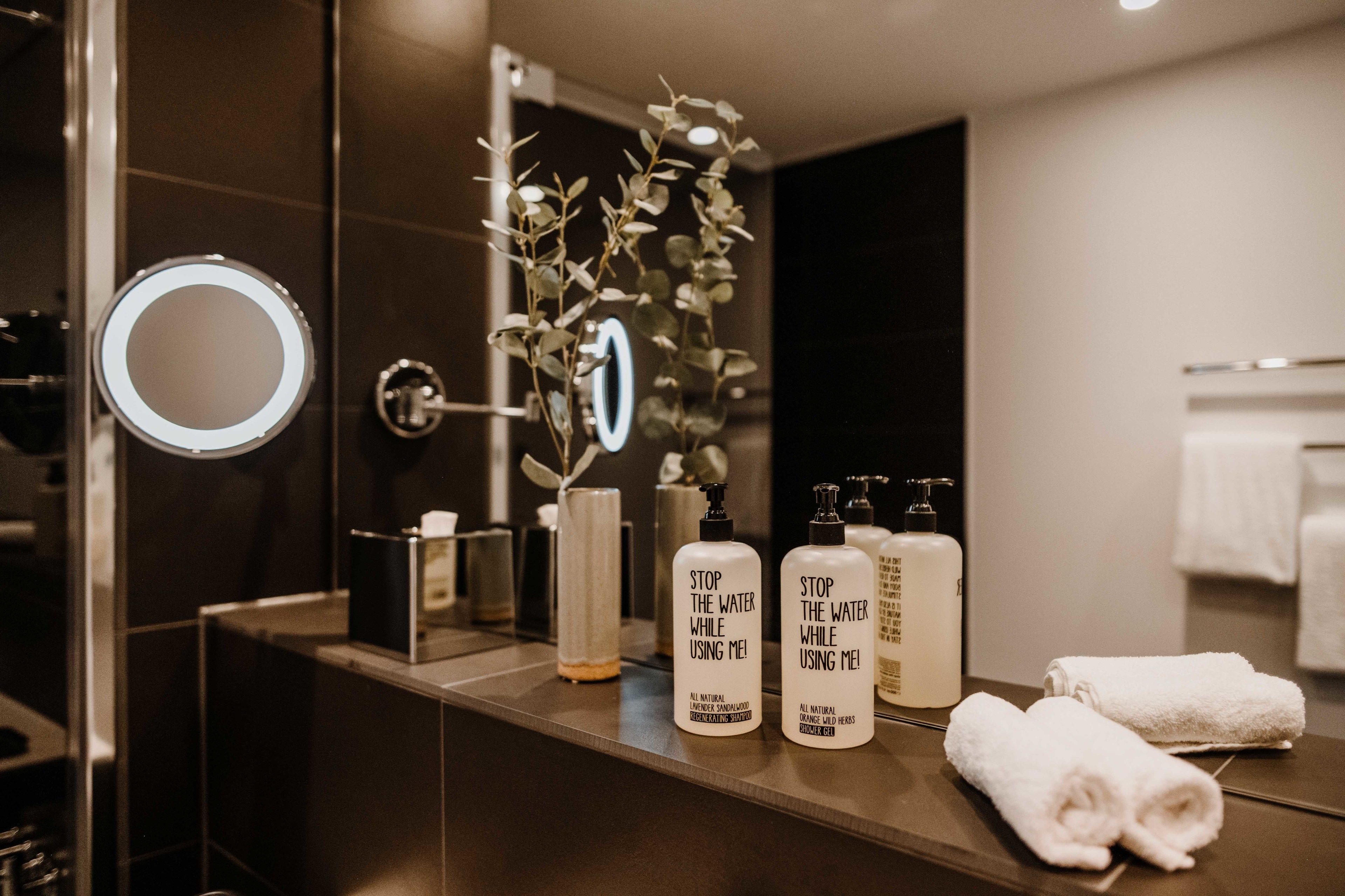A bathroom with a mirror, two bottles of 'Stop the Water While Using Me!' shampoo, a plant, and two rolled towels on the counter.