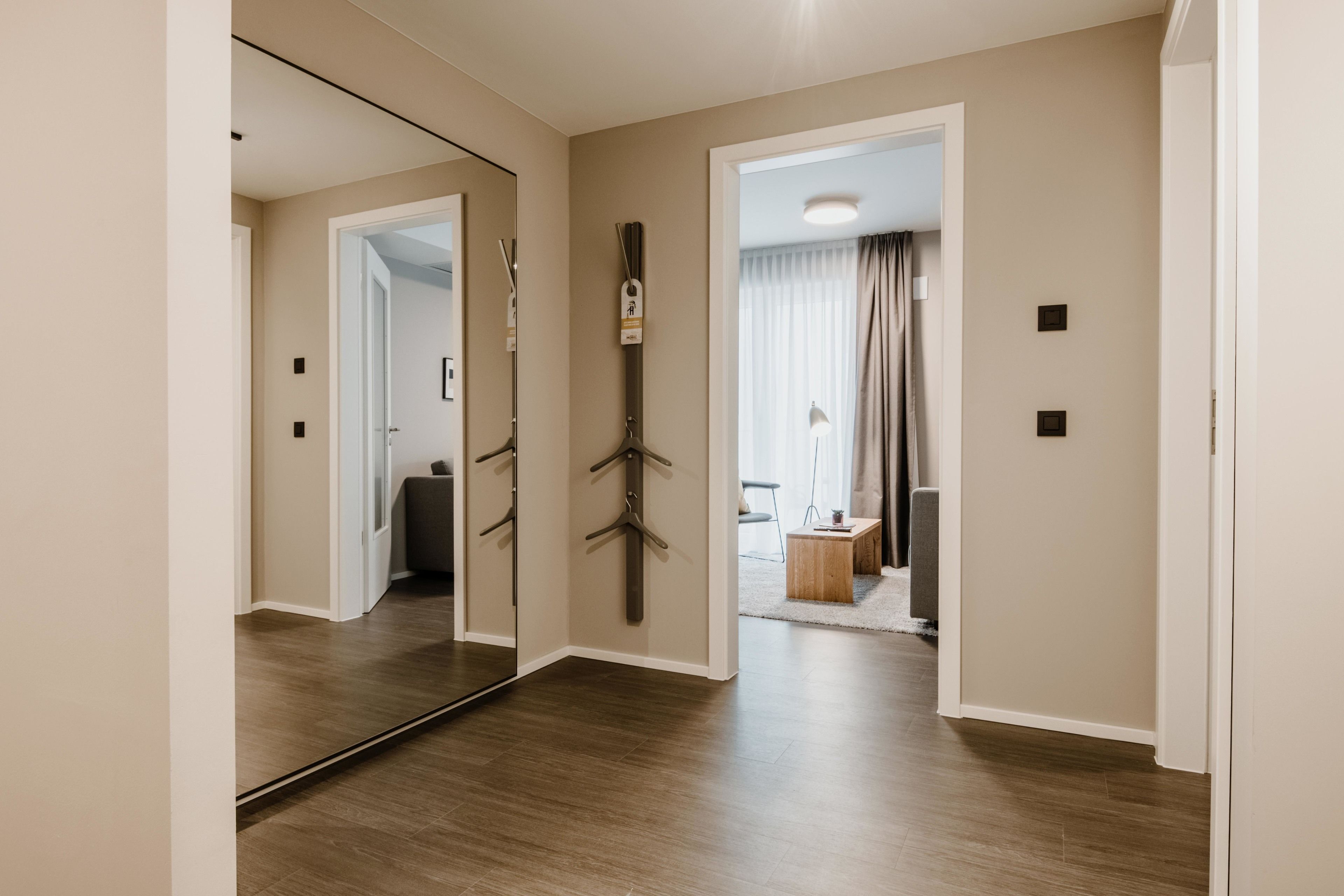 A hallway with a large mirror, wooden flooring, and a door leading to a room with a bed and curtains.