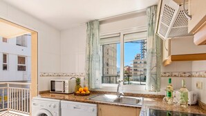 A compact kitchen with a washing machine, a microwave, a sink, and a small island with a stove. - Apartment Arenal 4 - PlusHolidays (Calpe)