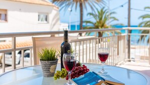 A table with a bottle of wine, two glasses of red wine, a book, and a potted plant on a balcony with a view of a palm tree and a building. - Apartment Arenal 4 - PlusHolidays (Calpe)