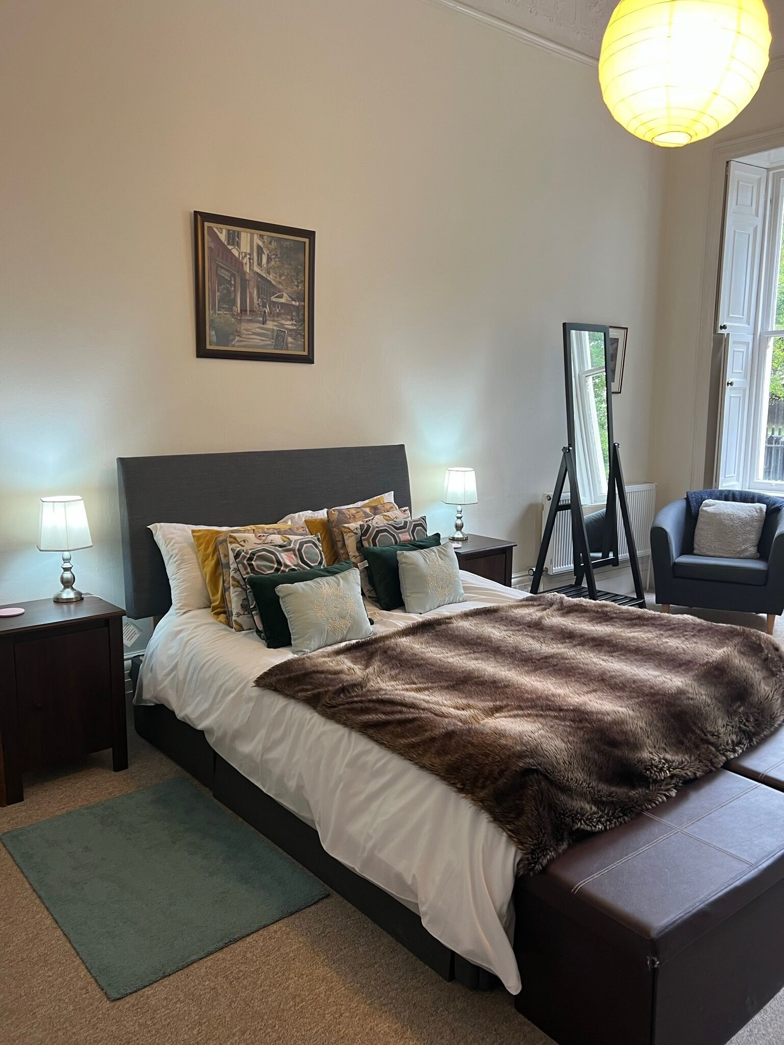 A neatly arranged bedroom with a large bed, a standing mirror, a chair, and a framed picture on the wall.