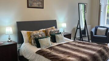 A neatly arranged bedroom with a large bed, a standing mirror, a chair, and a framed picture on the wall.