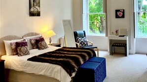 Ein Schlafzimmer mit einem großen Bett, einem Schreibtisch und einem Stuhl. Fenster bieten Blick auf Grünflächen außerhalb.