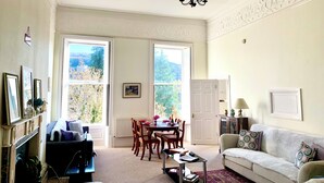 Ein Wohnzimmer mit Kamin, einer Couch, einem Esstisch mit Stühlen und großen Fenstern mit Blick auf Bäume und Hügel.