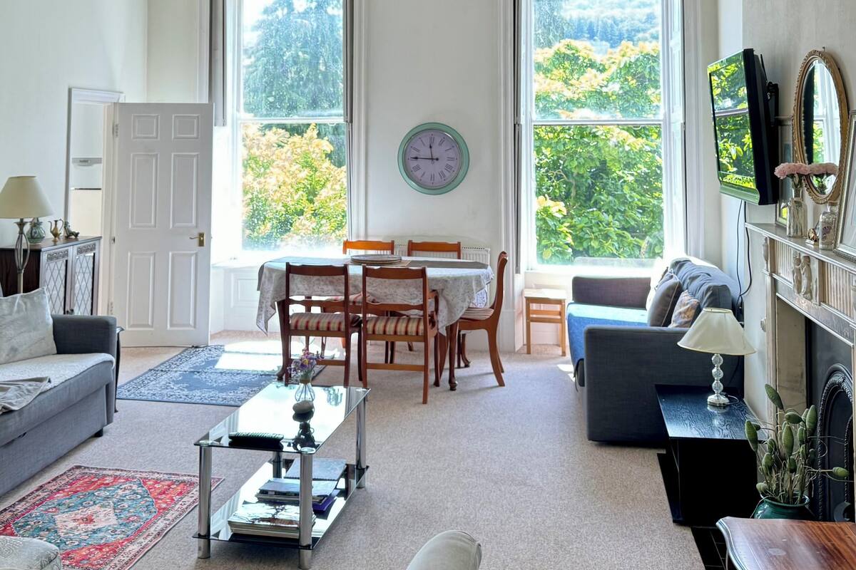 Uma sala de estar espaçosa com área de jantar, janelas amplas com vista para o verde, um lustre e um relógio na parede.