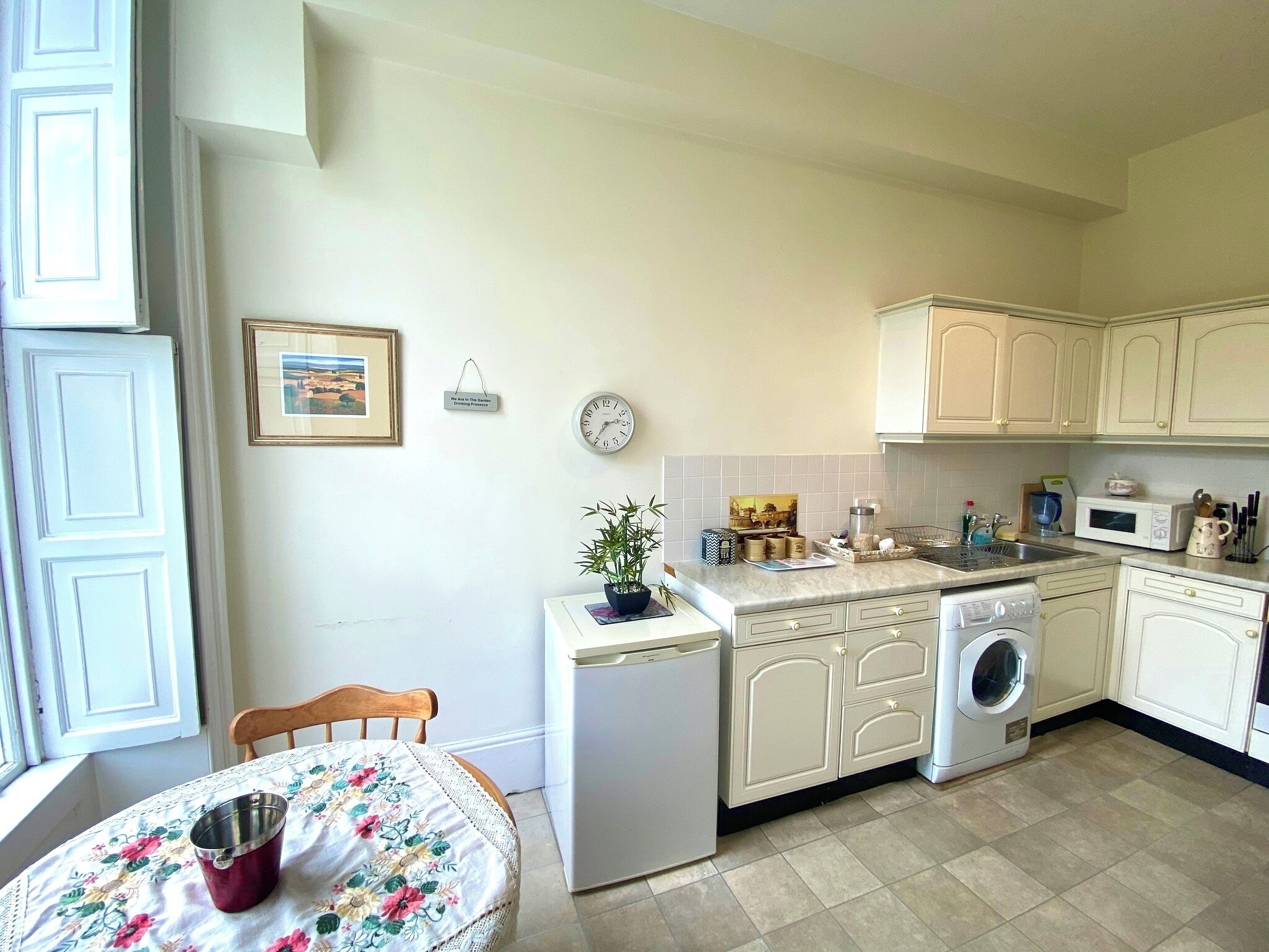 A compact kitchen with a sink, microwave, and a small refrigerator. There is a dining table with a floral tablecloth and a chair.
