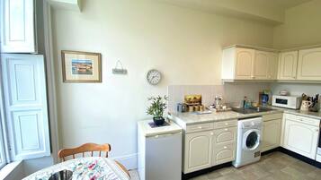 A compact kitchen with a sink, microwave, and a small refrigerator. There is a dining table with a floral tablecloth and a chair.