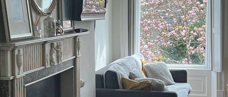 A living room with a fireplace, a sofa, a television, and a window with a view of blooming trees.