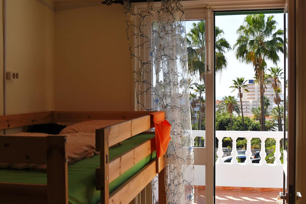 A room with a wooden bed, a balcony with a white railing, and a view of palm trees and buildings.