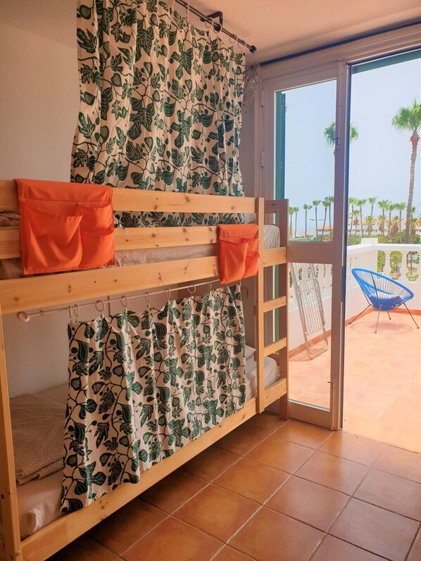 Una cama individual con cortinas estampadas con hojas, un banco de madera con cojines anaranjados, y vistas a palmeras y una silla afuera.