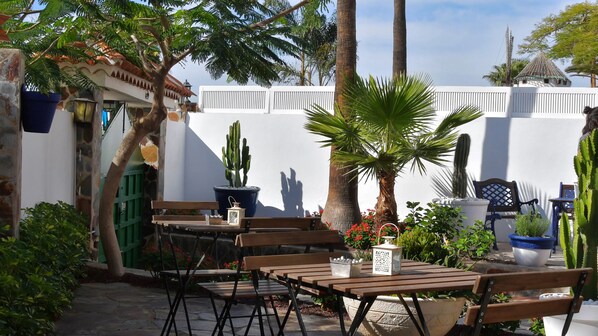 An outdoor patio with wooden tables and chairs, surrounded by potted plants and cacti.