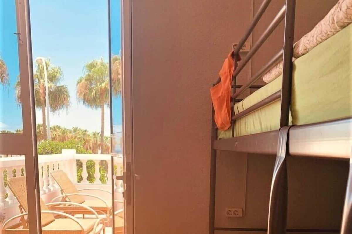 A room with bunk beds, a sliding glass door leading to an outdoor area with chairs and a view of palm trees.
