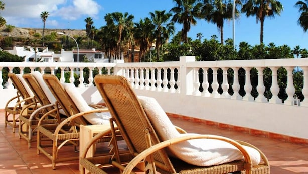 Filas de sillones de mimbre con cojines en un patio empedrado, con vistas a un paisaje costero con palmeras y un cielo azul despejado.