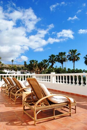 A row of wicker lounge chairs with cushions on a tiled patio, overlooking a coastal landscape with palm trees and a clear blue sky.