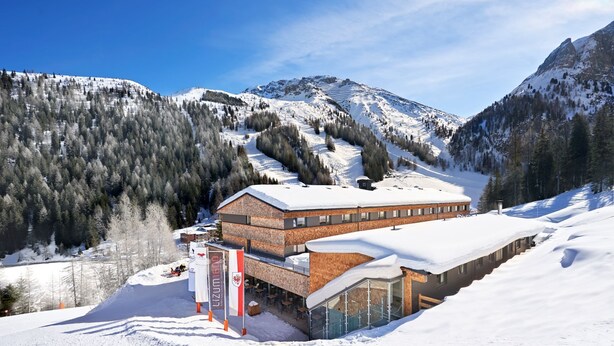 A modern ski resort building with a glass facade, surrounded by snow-covered mountains and trees.