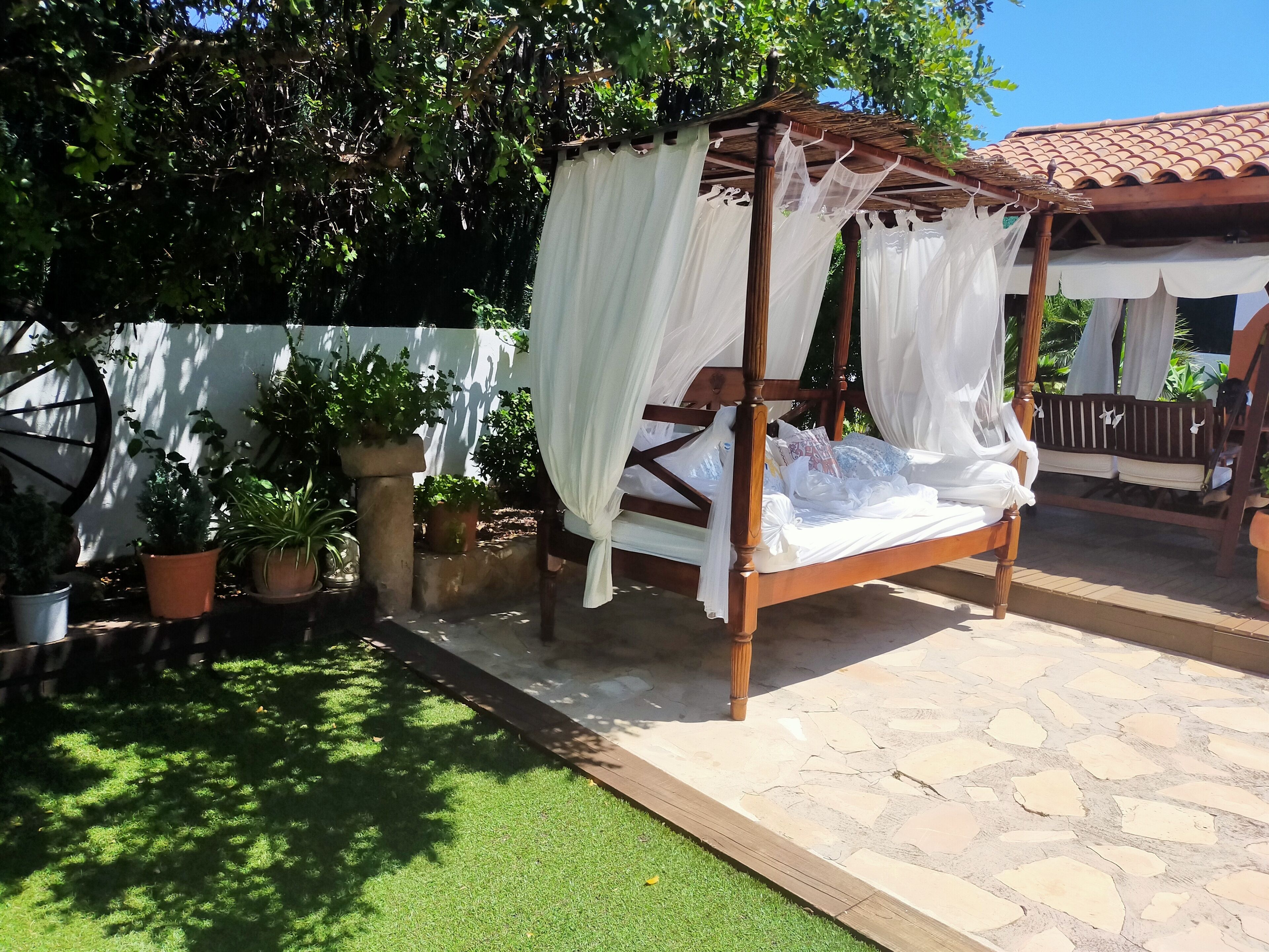 Ein hölzernes Himmelbett mit weißen Vorhängen, umgeben von Topfpflanzen und einer steinbelagten Terrasse.
