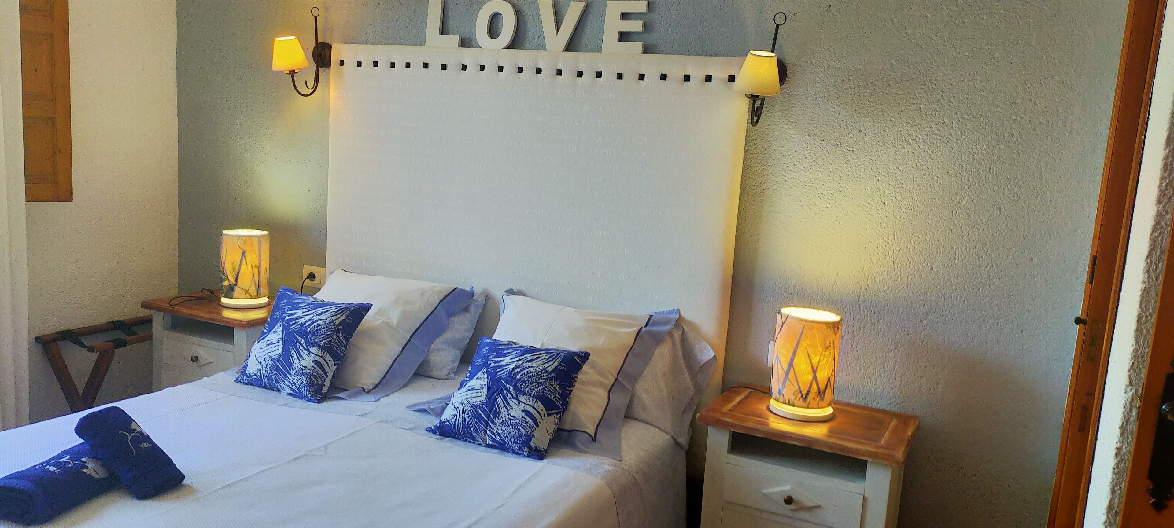 Un lit bien fait, avec du linge de lit blanc et des coussins à motifs bleus, flanqué de tables de chevet sur lesquelles sont posées des lampes, et un panneau mural sur lequel on peut lire le mot « AMOUR ».