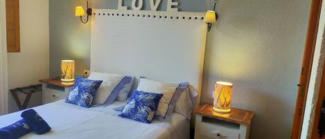 Un lit bien fait, avec du linge de lit blanc et des coussins à motifs bleus, flanqué de tables de chevet sur lesquelles sont posées des lampes, et un panneau mural sur lequel on peut lire le mot « AMOUR ».