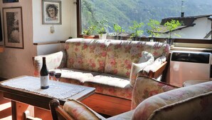 A cozy room with a floral sofa, a table with a bottle and glasses, and a view of mountains through a large window. - B&B Rocca di Bajedo (Pasturo)