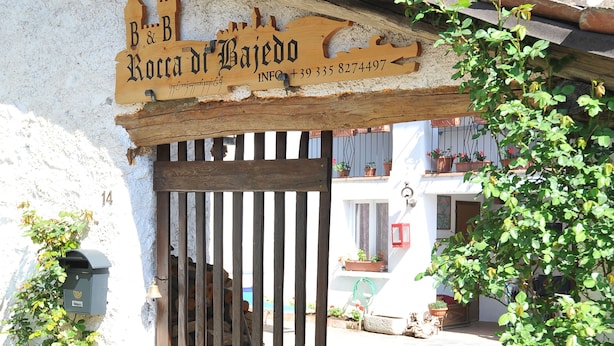 A rustic wooden gate with a sign above it, a white building with a tiled roof, and potted plants on the balcony. - B&B Rocca di Bajedo (Pasturo)