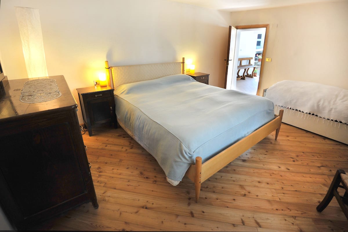 A bedroom with a wooden floor, a bed with a light blue bedspread, a bedside table with a lamp, and a wooden dresser.