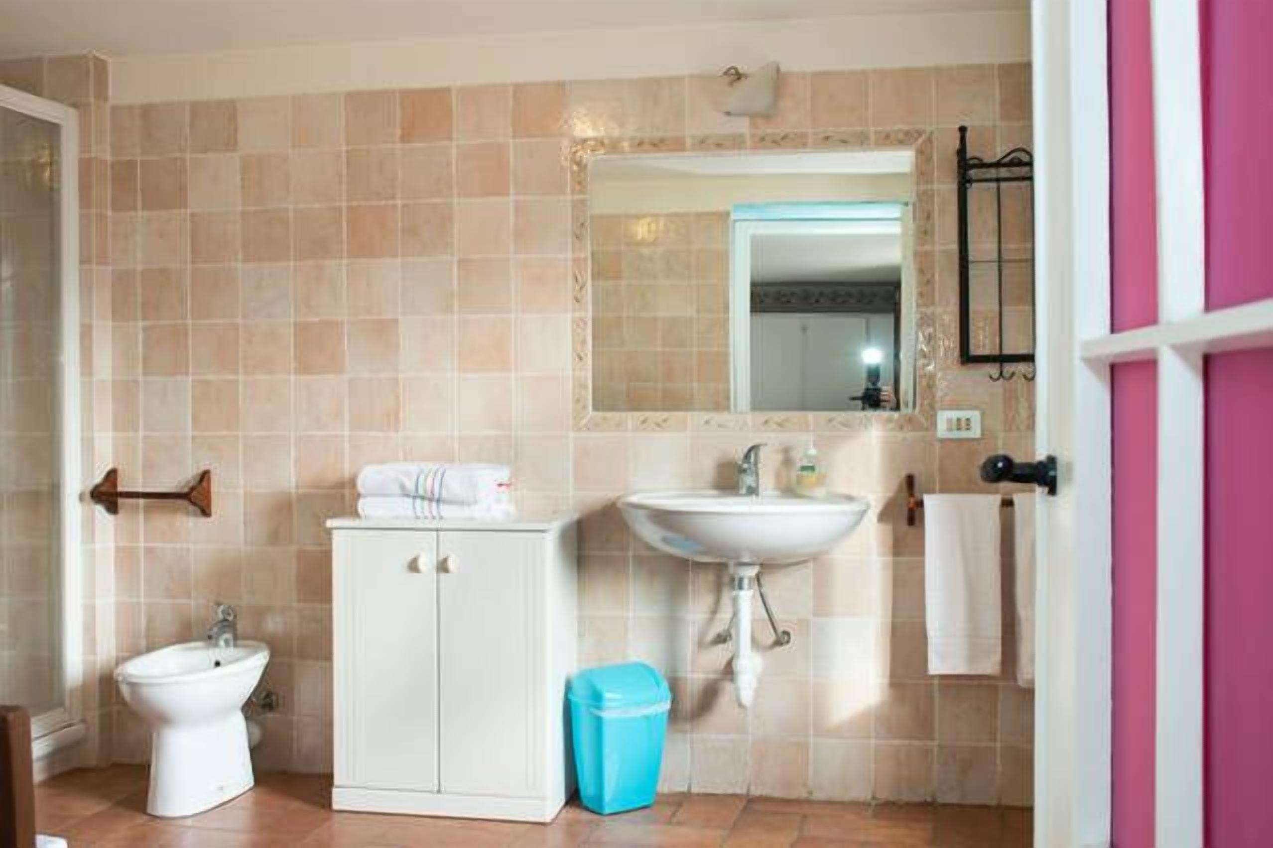 Et badeværelse med beige fliser, en hvid vask, en bidet, et skab og en blå skraldespand.