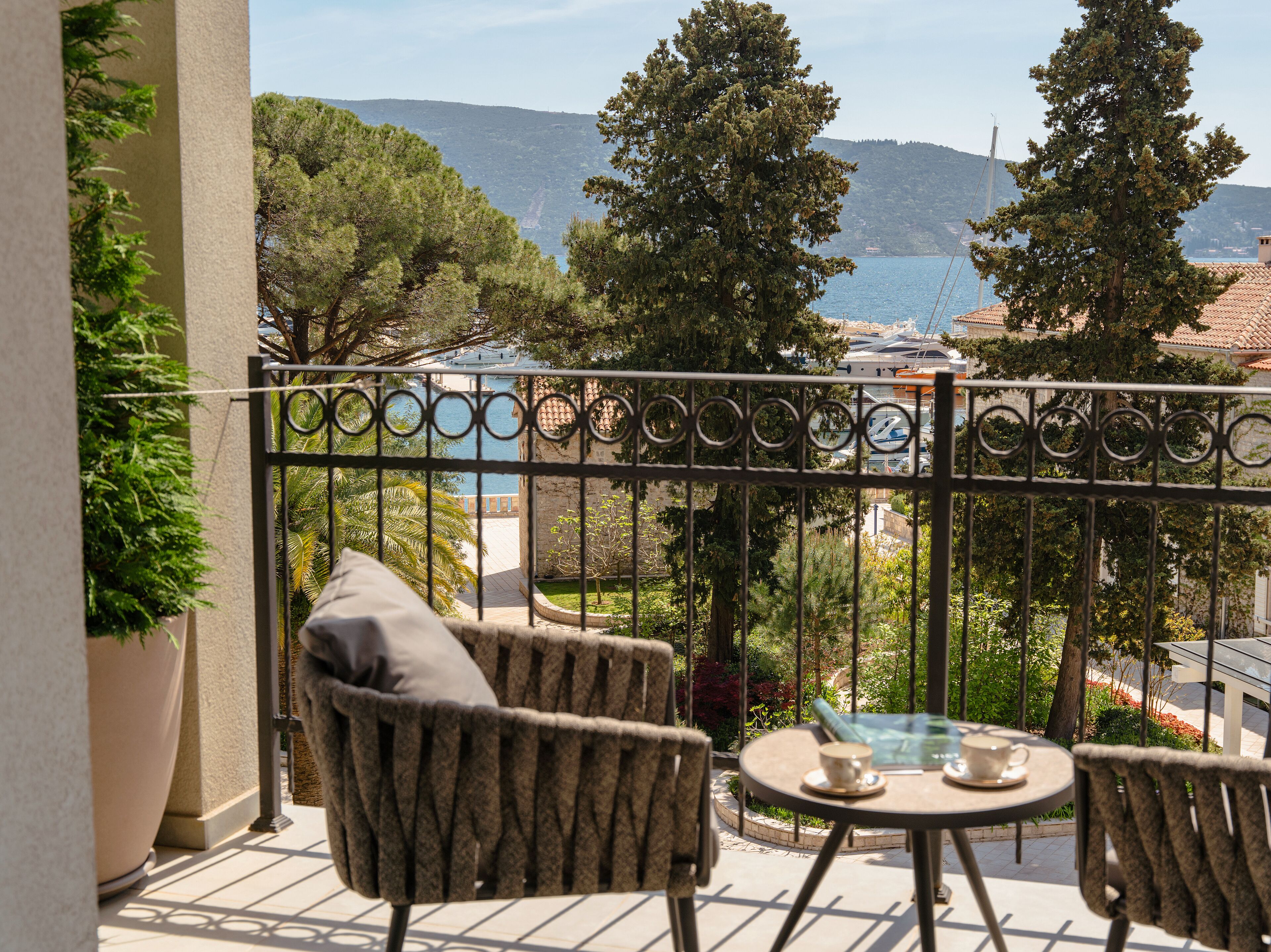 A balcony with wicker furniture, a table with a cup and saucer, and a view of the sea and hills.