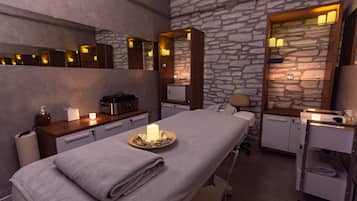Experience a serene atmosphere in this spa room with a massage table, candlelit ambiance, and a stone wall backdrop.