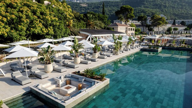 Un espace aménagé au bord de la piscine, avec des chaises longues et des parasols, une piscine à eau claire et une vue sur une colline parsemée de bâtiments.