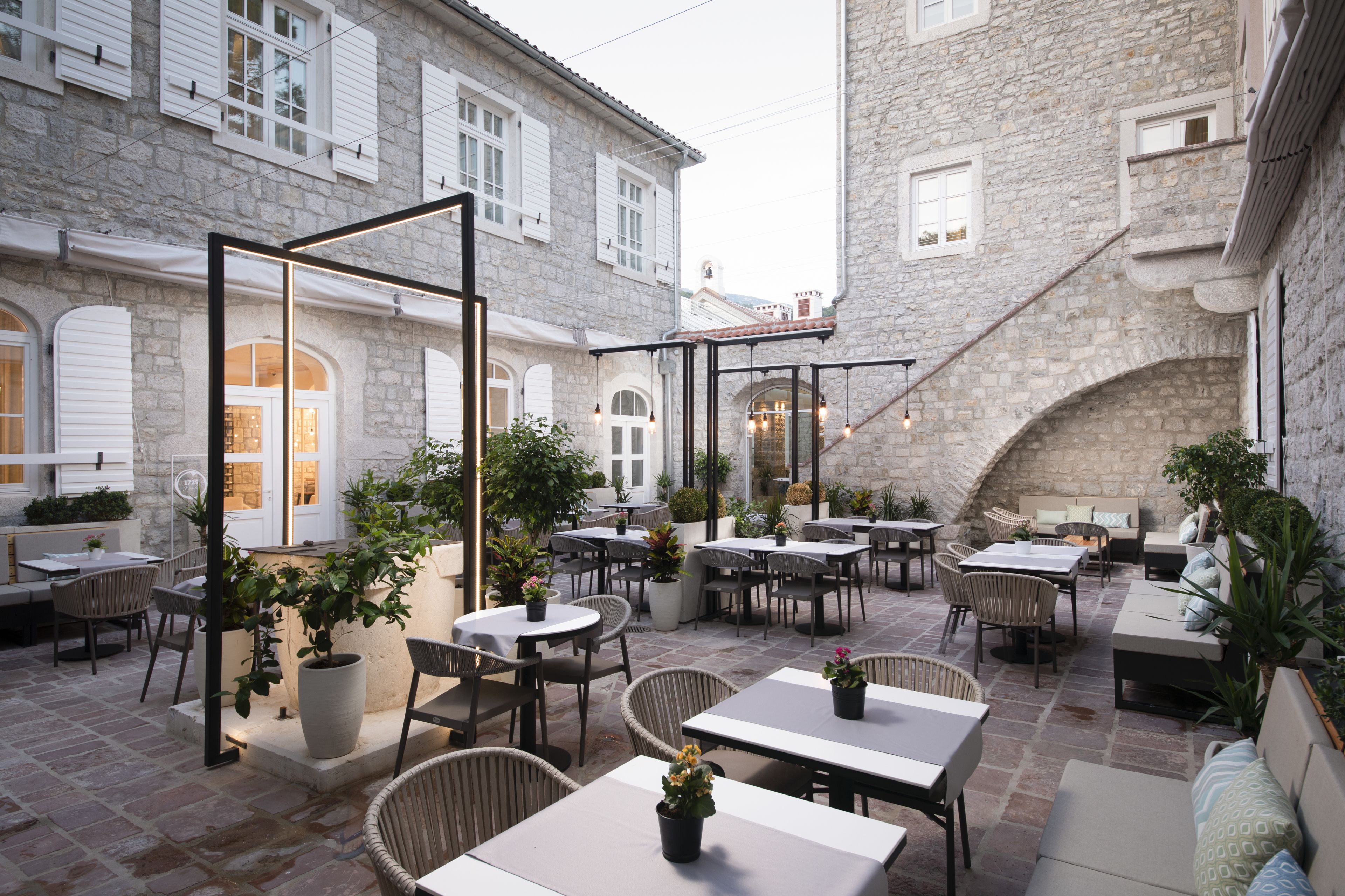 An outdoor café with stone buildings, tables, chairs, and potted plants.