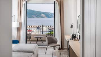 Habitación de hotel con balcón, una cama, una silla, un escritorio y vistas al mar y a las colinas.