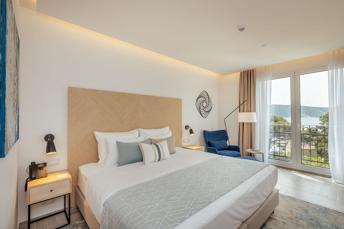 Ein modernes Hotelzimmer mit einem großen Bett, einem Schreibtisch, einem Sessel und Blick ins Freie, sowie einer Wanddekoration.