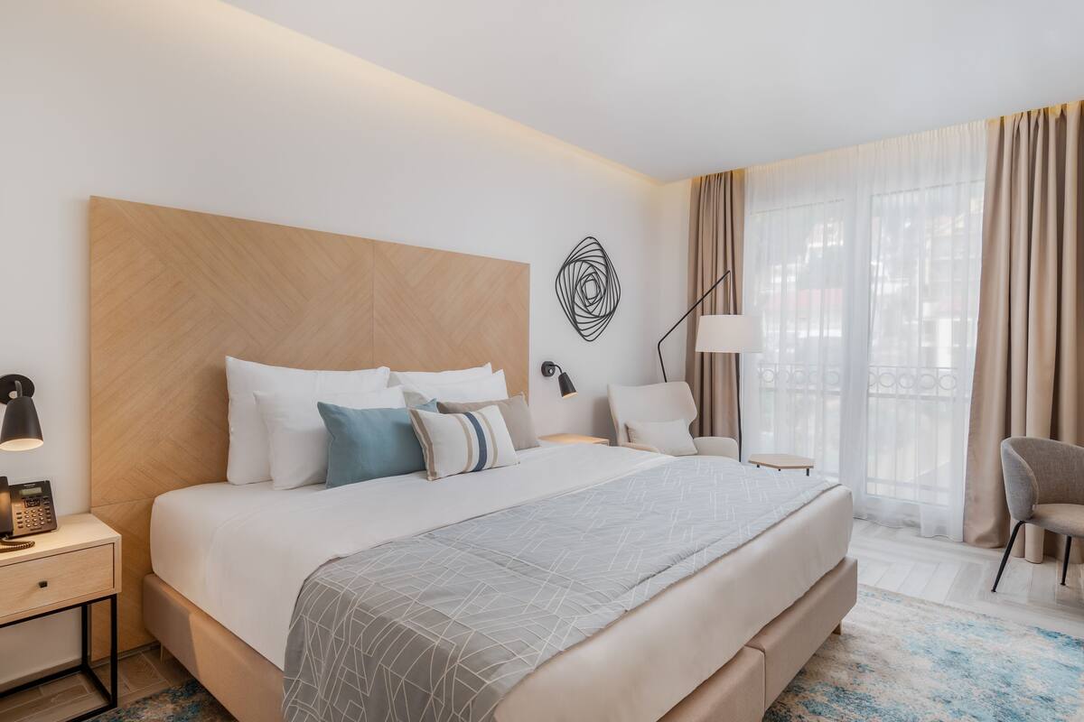 Ein Hotelzimmer mit einem großen Bett, einem Nachttisch mit Lampe, einem Sessel und Blick durch ein mit Vorhängen drapiertes Fenster nach draußen.
